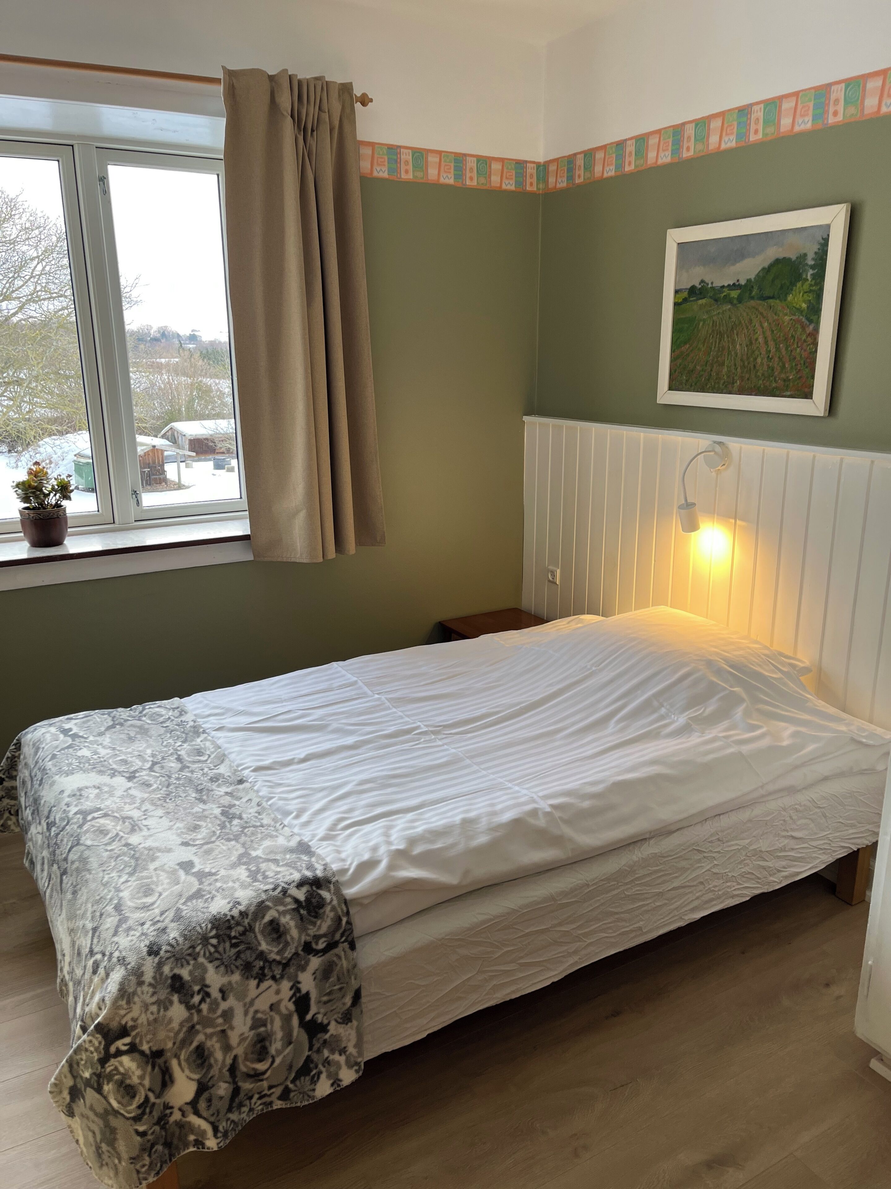 Quarto com cama, janela com cortinas, radiador, quadro na parede e uma pequena planta em vaso.
