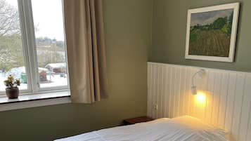 A bedroom with a bed, a window with curtains, a radiator, a wall-mounted picture, and a small potted plant.