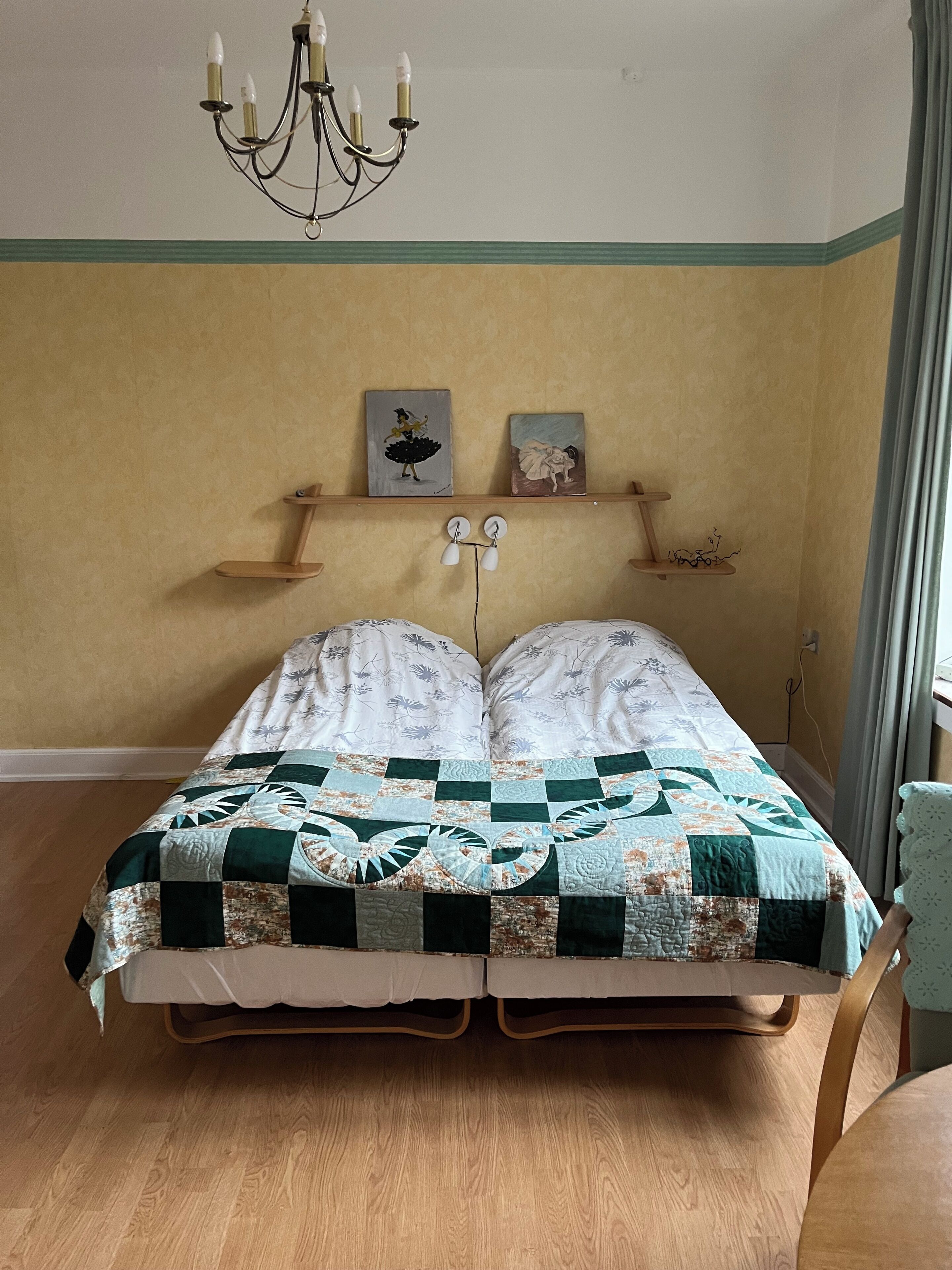 A bed with a quilt, a chandelier, and two paintings on the wall.