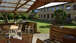 Ein sonniger Außenbereich mit Holztischen und -stühlen, einer überdachten Sitzgelegenheit und Blick auf ein Backsteingebäude im Hintergrund.