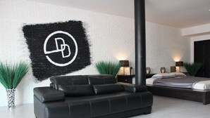 Une chambre moderne avec un canapé en cuir noir, un lit doté d’une tête de lit sombre et un mur en briques blanches orné d’un logo noir et blanc.
