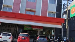 Un edificio de varios pisos con fachada roja y blanca, un letrero verde y coches y motocicletas estacionados adelante.