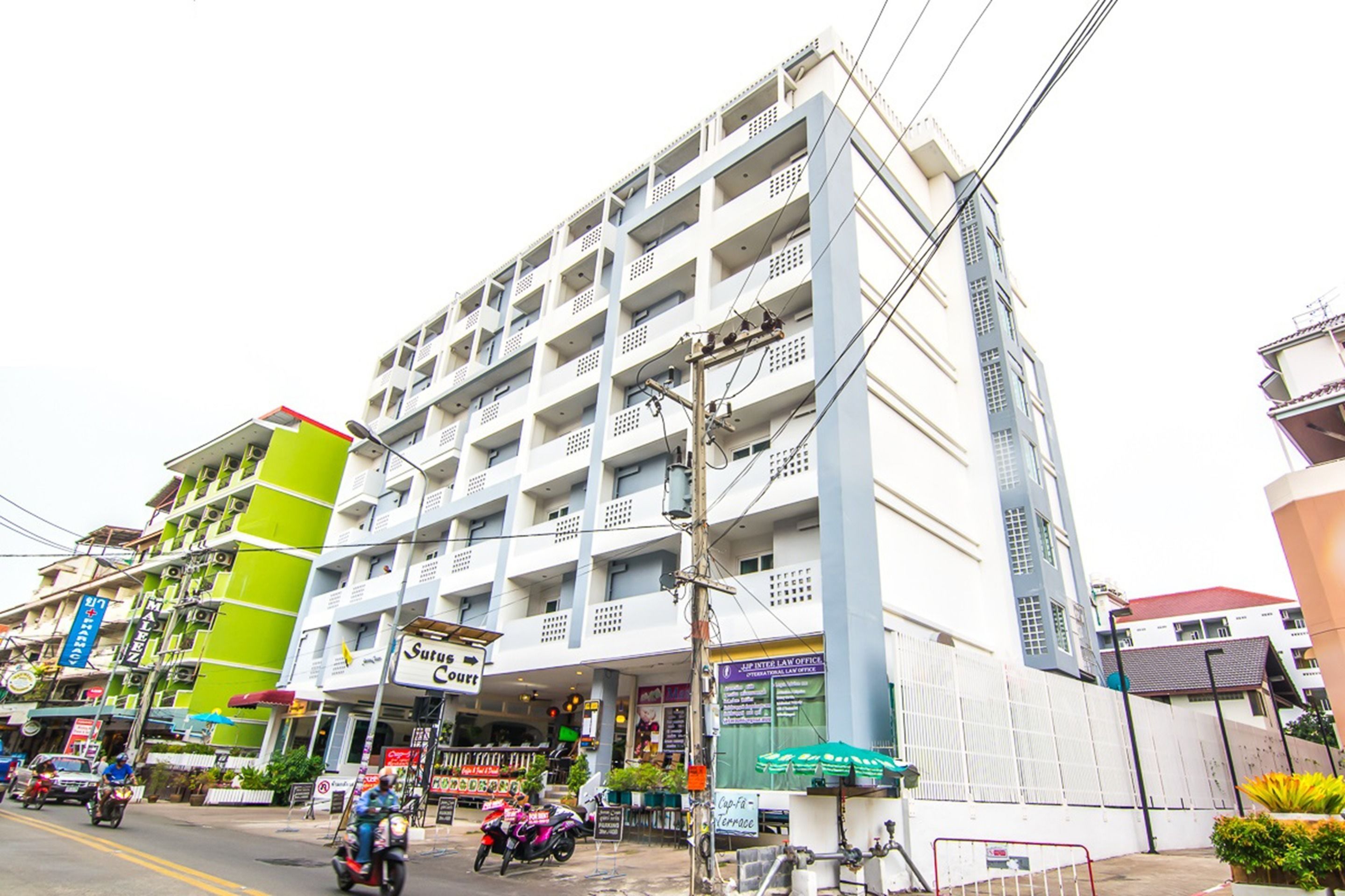 A multi-story hotel with balconies, a green building adjacent to it, and a street with motorcycles.
