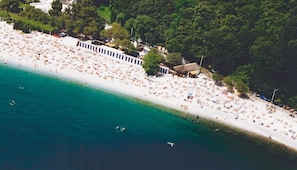 An aerial view of a beach with numerous sunbathers, a row of beach huts, and a dense forest in the background. - Hotel Ai Sette Nani (Duino-Aurisina)