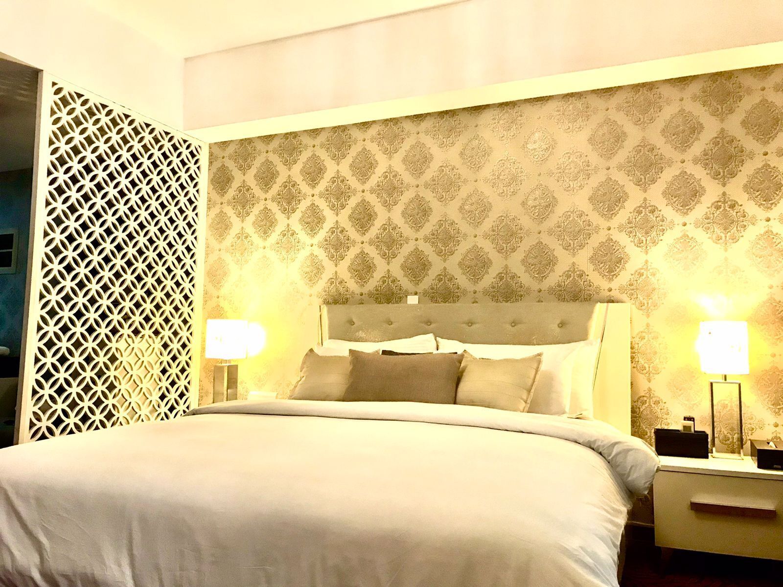 A neatly made bed with a tufted headboard, two bedside lamps, and a patterned wallpaper.