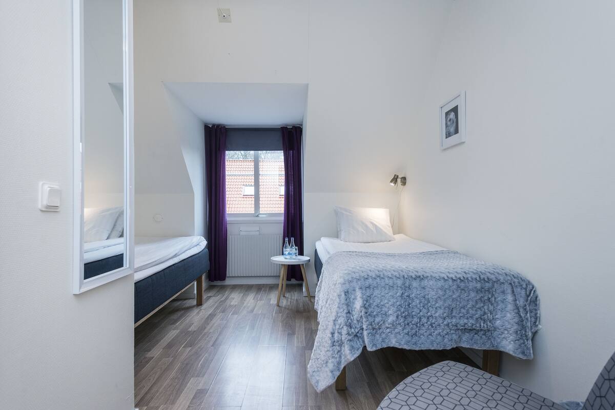 A small, well-lit room with a bed, a chair, a small table with a water bottle, and a window with curtains.