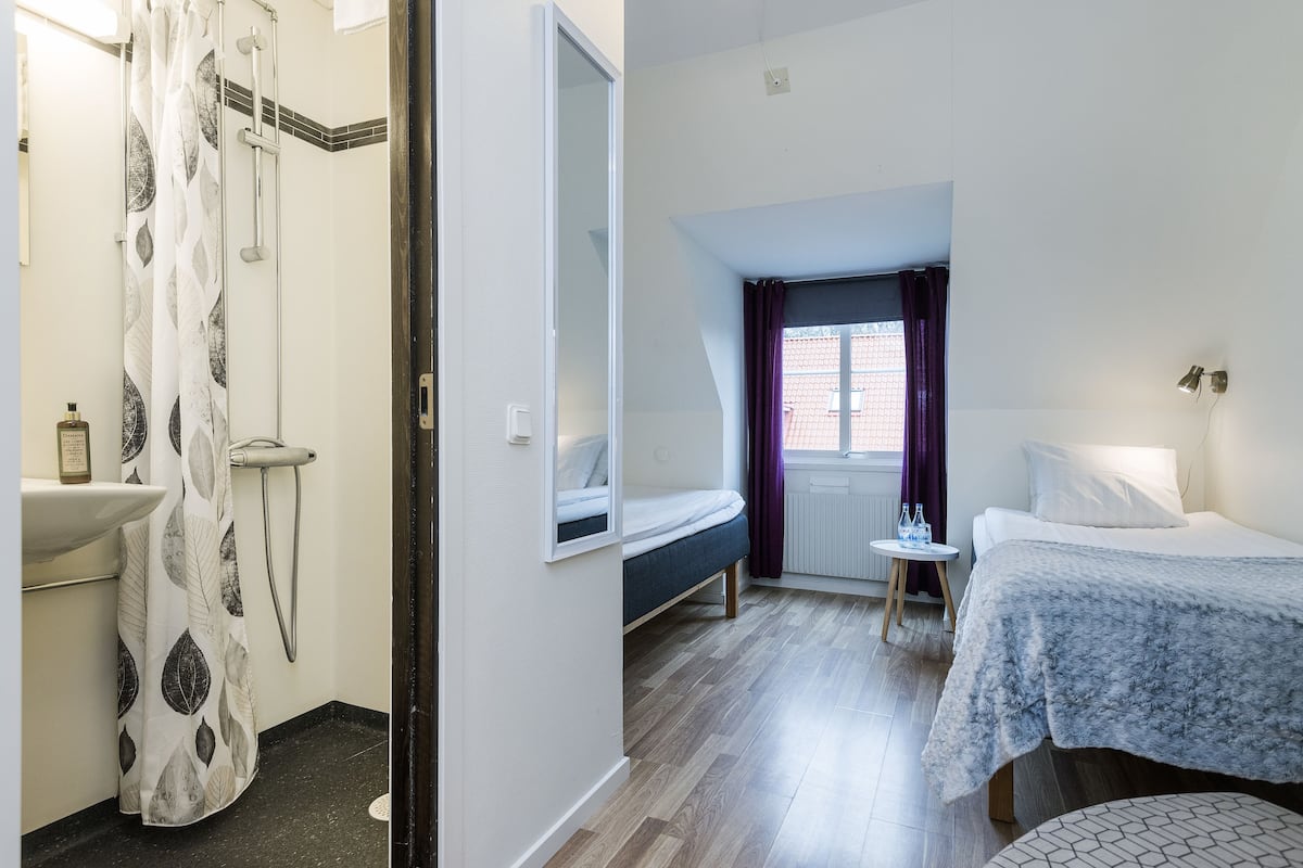 A compact hotel room with a shower, a bed, a bedside table with a lamp, and a window with curtains.