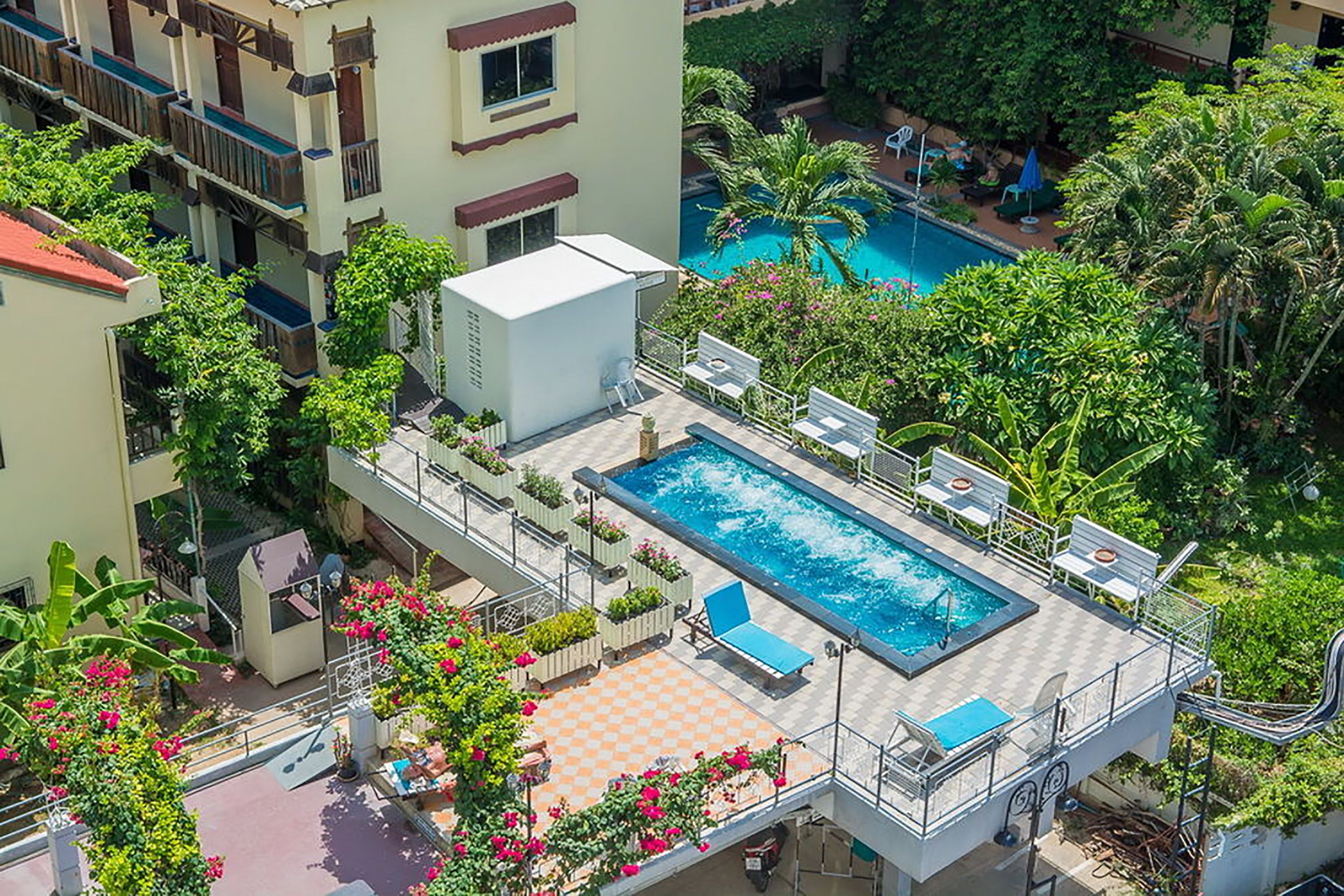 A rooftop swimming pool with lounge chairs and a checkered deck area, surrounded by lush greenery and a multi-story building with balconies.