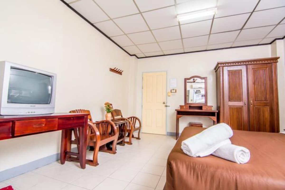 A hotel room with a bed, a desk with a television, a chair, a wooden wardrobe, and a door.