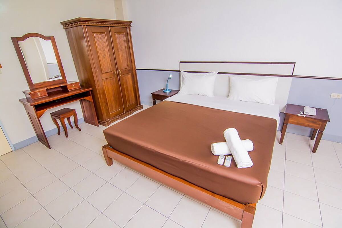A hotel room with a large bed, wooden wardrobe, a desk with a mirror, and a bedside table with a lamp.