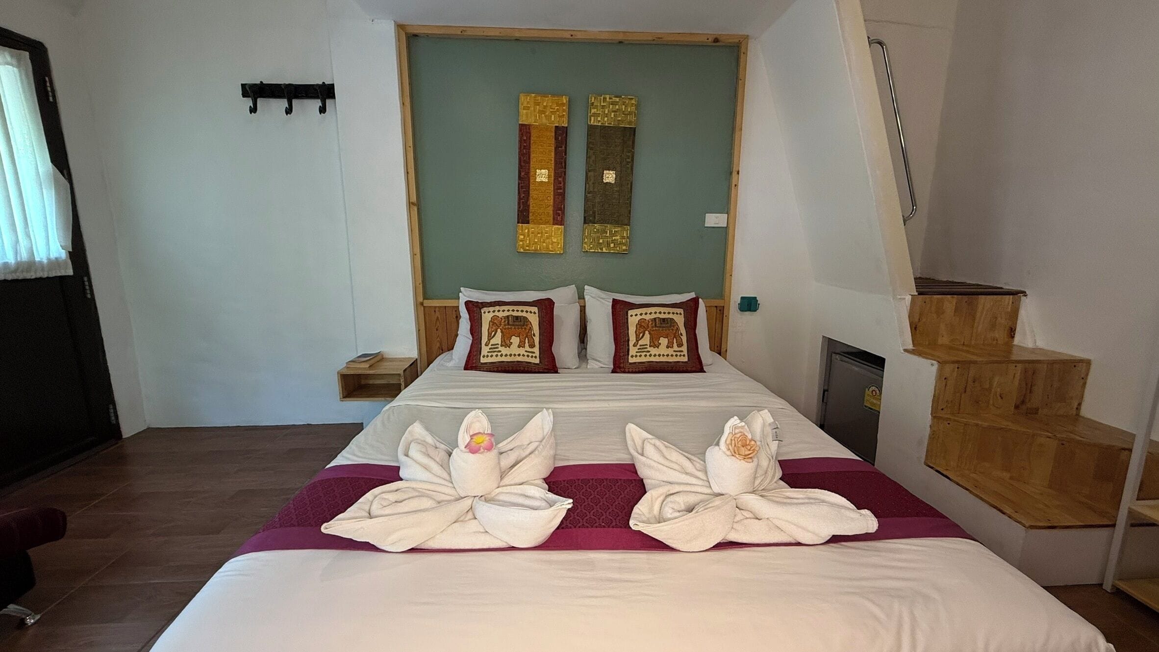 Une chambre d’hôtel avec un grand lit, deux oreillers ornés de motifs d’éléphants, une tête de lit en bois, une petite table de chevet et un escalier en bois.
