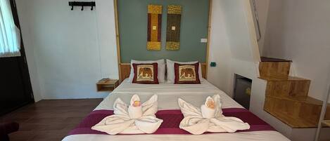 Une chambre d’hôtel avec un grand lit, deux oreillers ornés de motifs d’éléphants, une tête de lit en bois, une petite table de chevet et un escalier en bois.