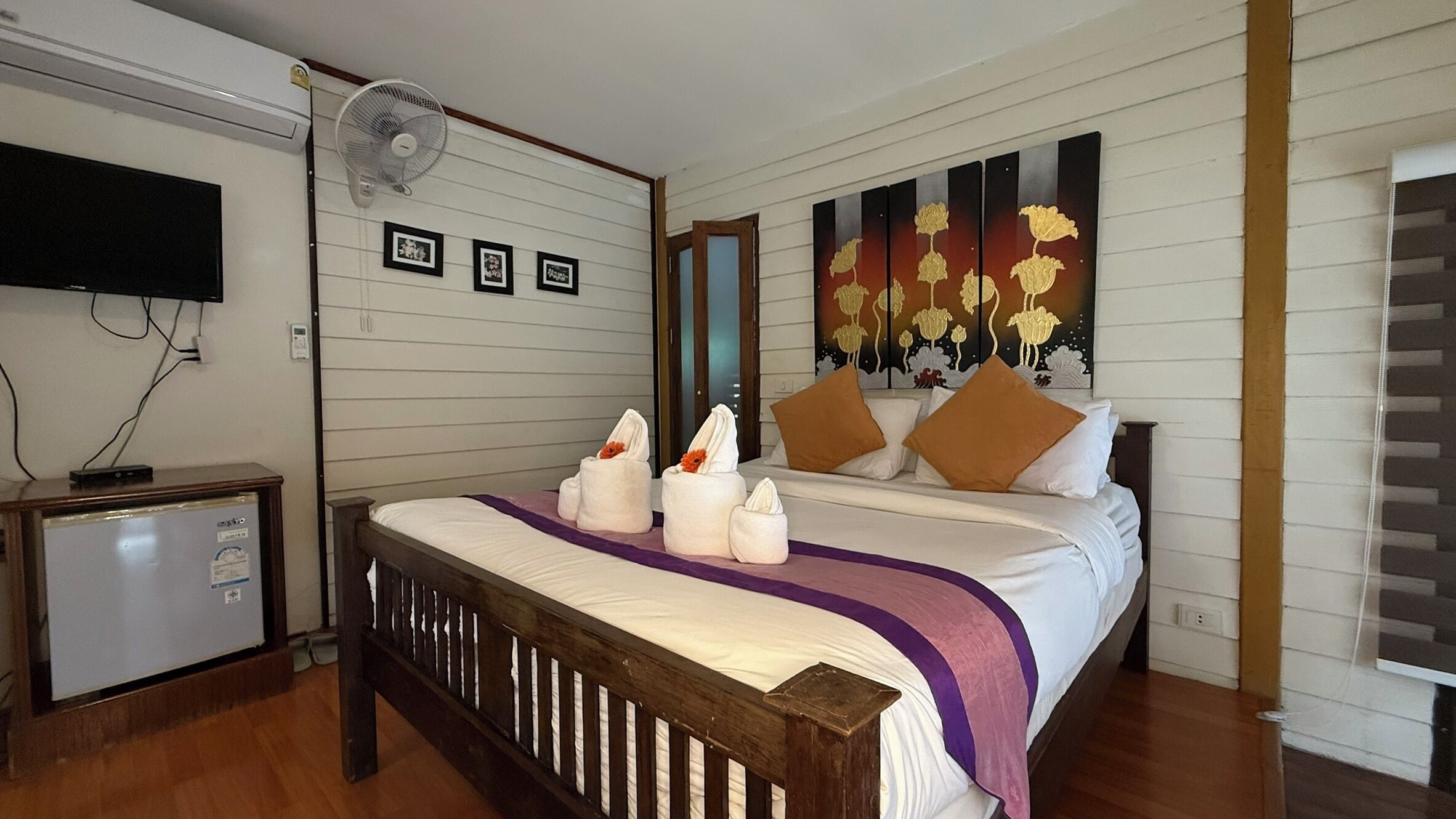 Une chambre d’hôtel avec un lit en bois, un petit bureau, un réfrigérateur, un téléviseur fixé au mur et un grand tableau représentant des fleurs.