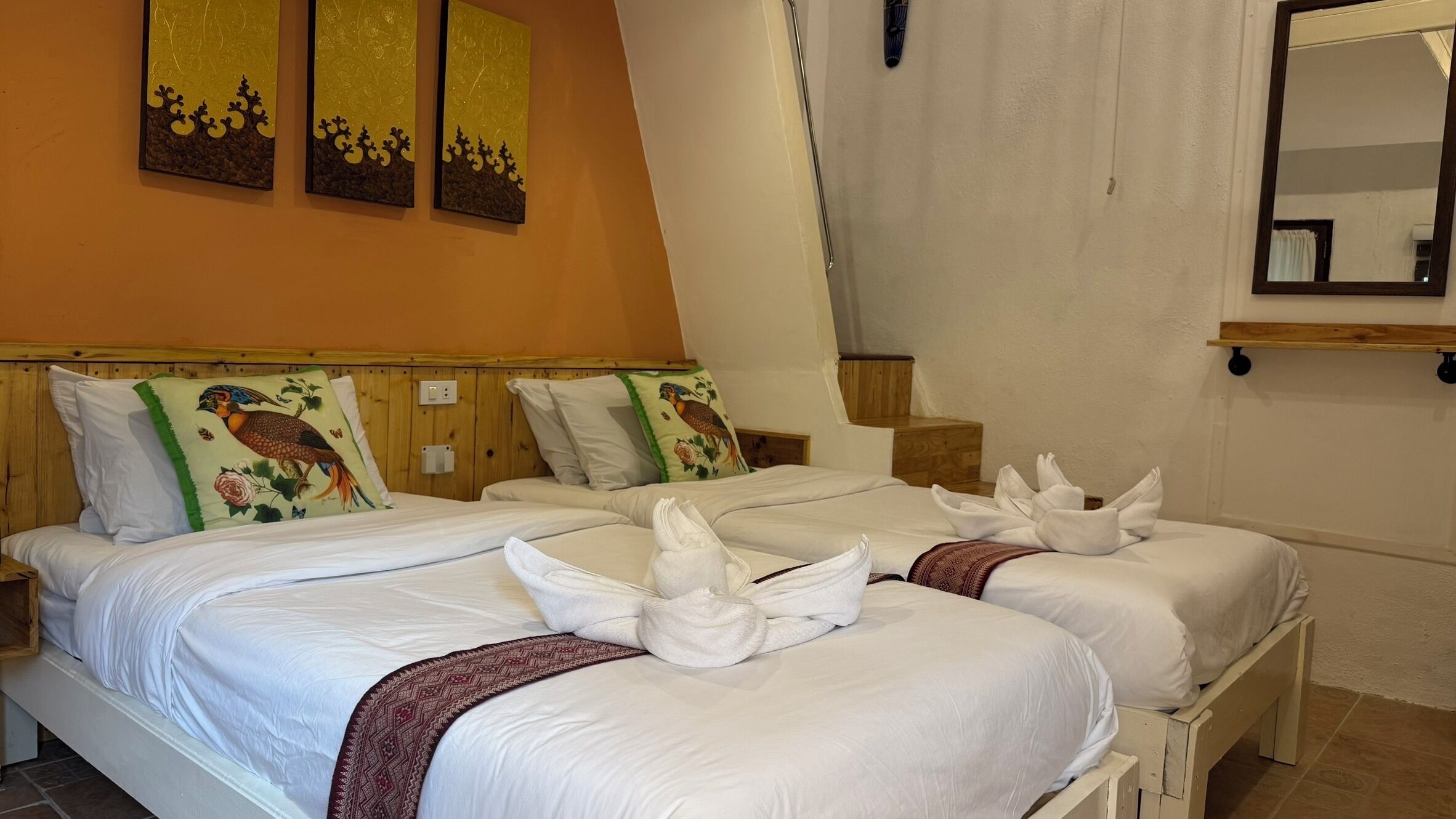 Habitación de hotel con dos camas, cada una con ropa blanca de cama y almohadas decorativas, un cabecero de madera y un espejo en la pared.
