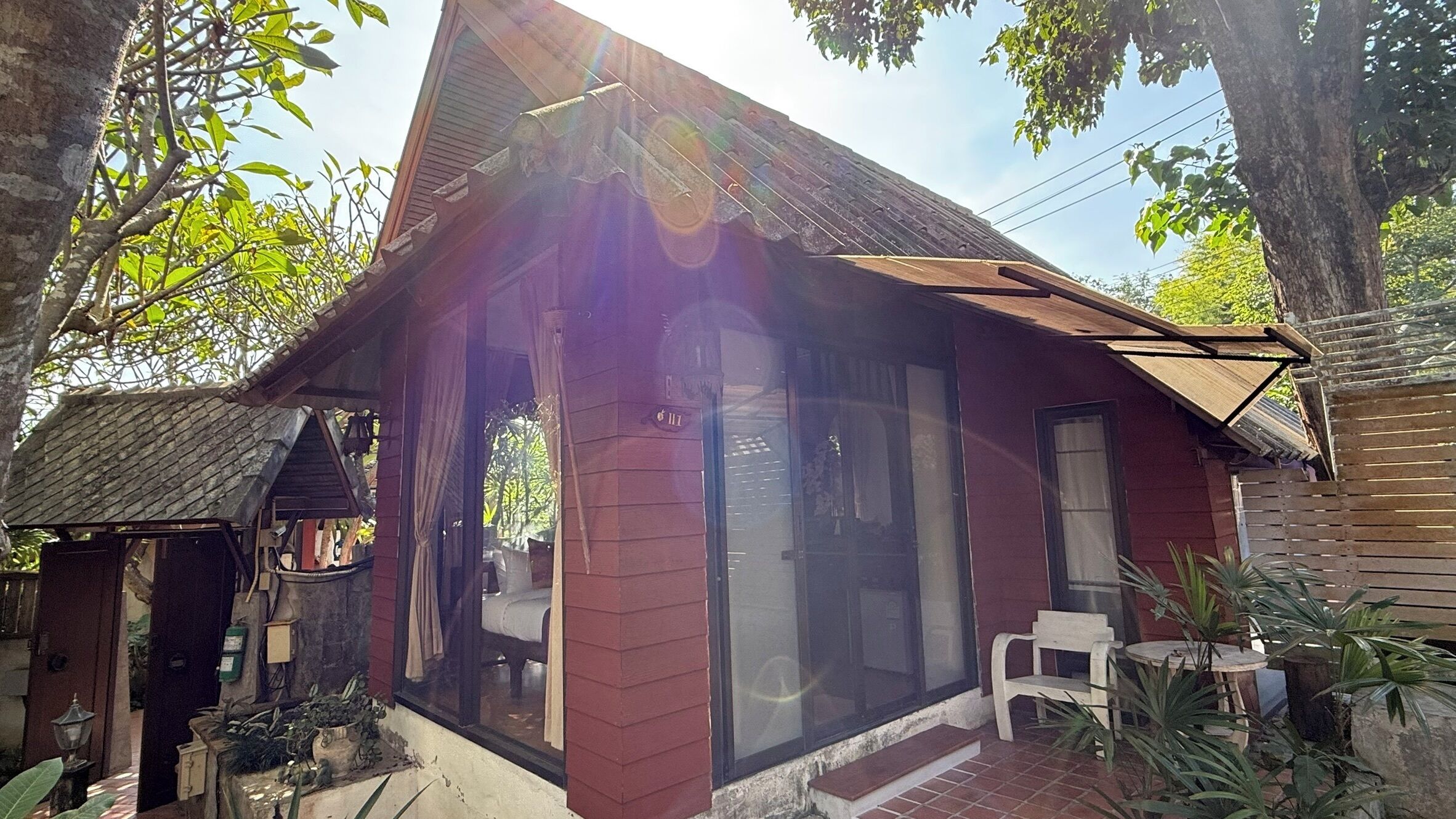 A rustic house with a red exterior, large glass windows, and a small porch area with a chair and table.
