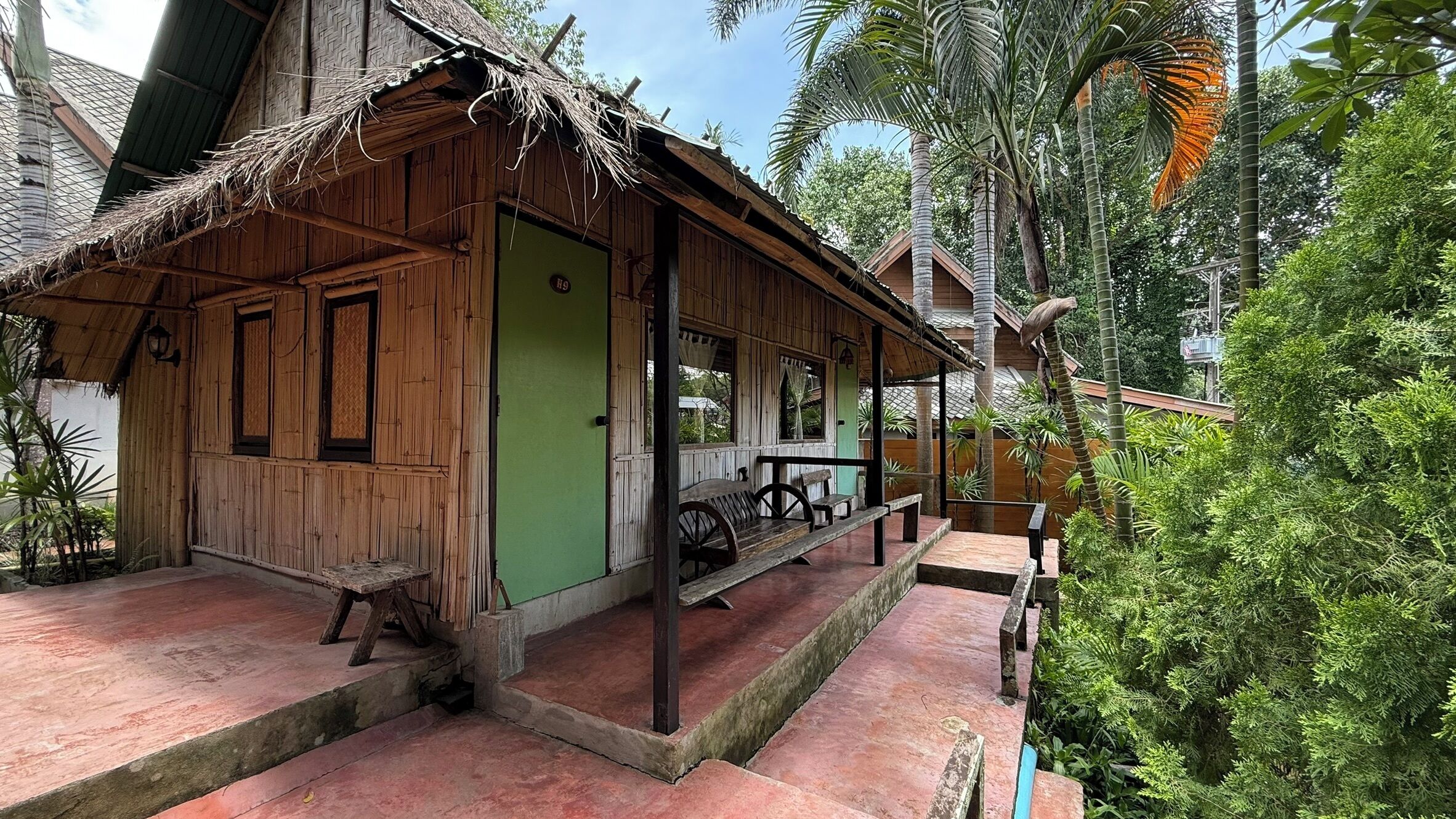 A traditional wooden house with a thatched roof, a porch with benches, and a green wall.