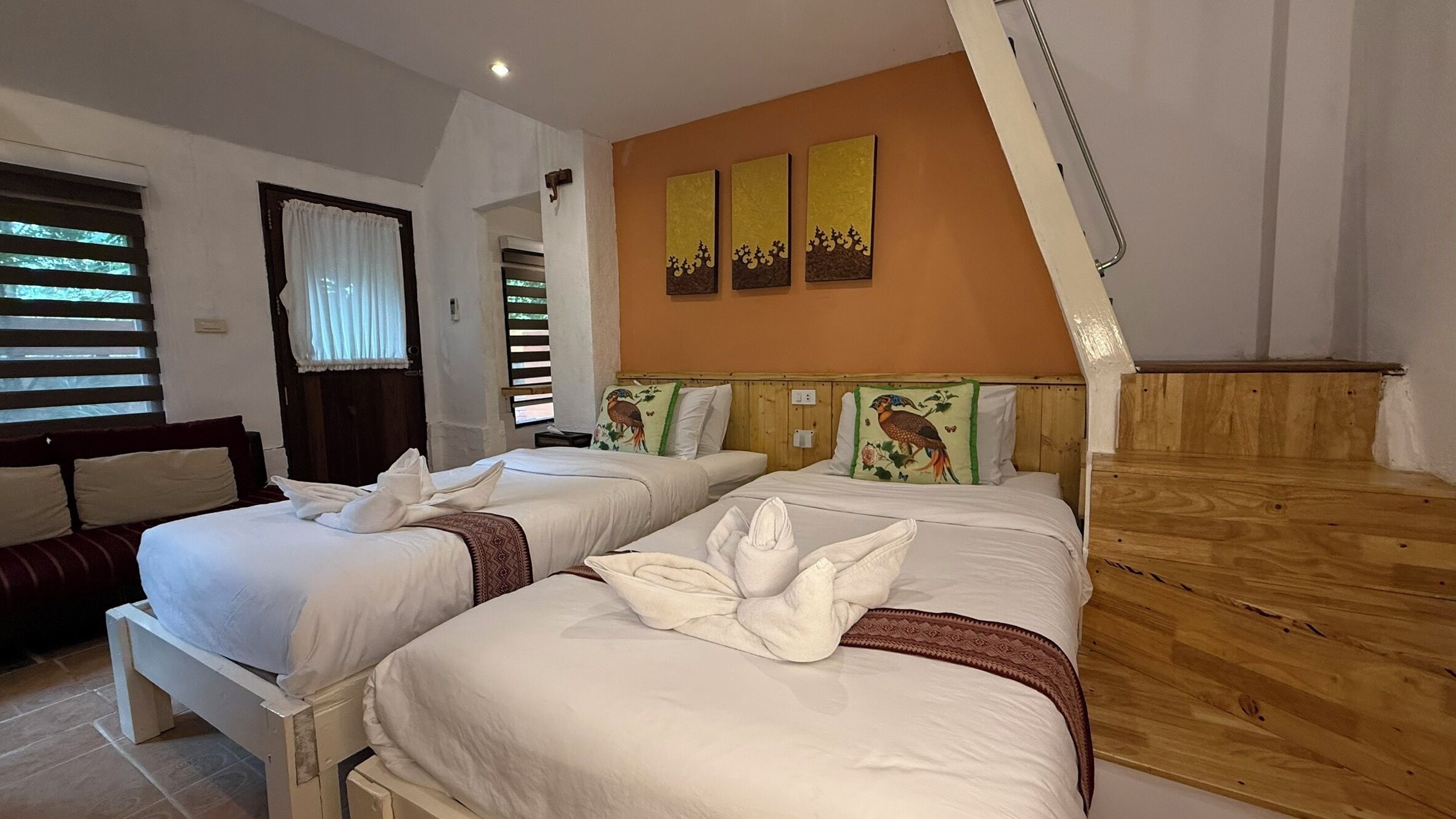 A hotel room with two beds, a wooden headboard, a window with blinds, and a wall with three paintings.