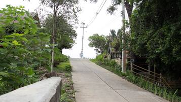 A paved pathway flanked by lush greenery and a rustic fence, leading towards a distant structure.