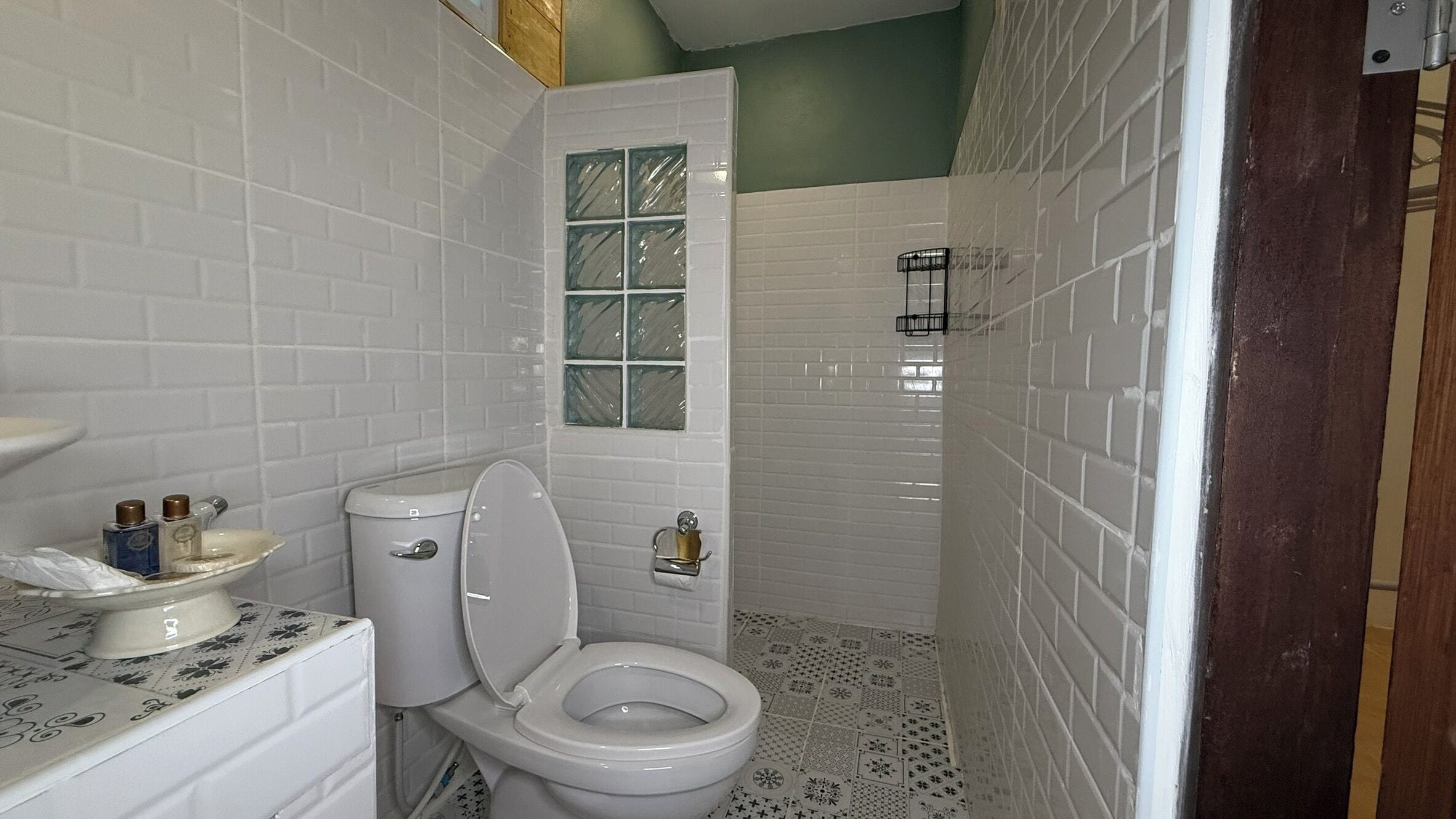 A bathroom with white subway tiles, a toilet with a glass window, a sink with decorative bottles, and patterned floor tiles.
