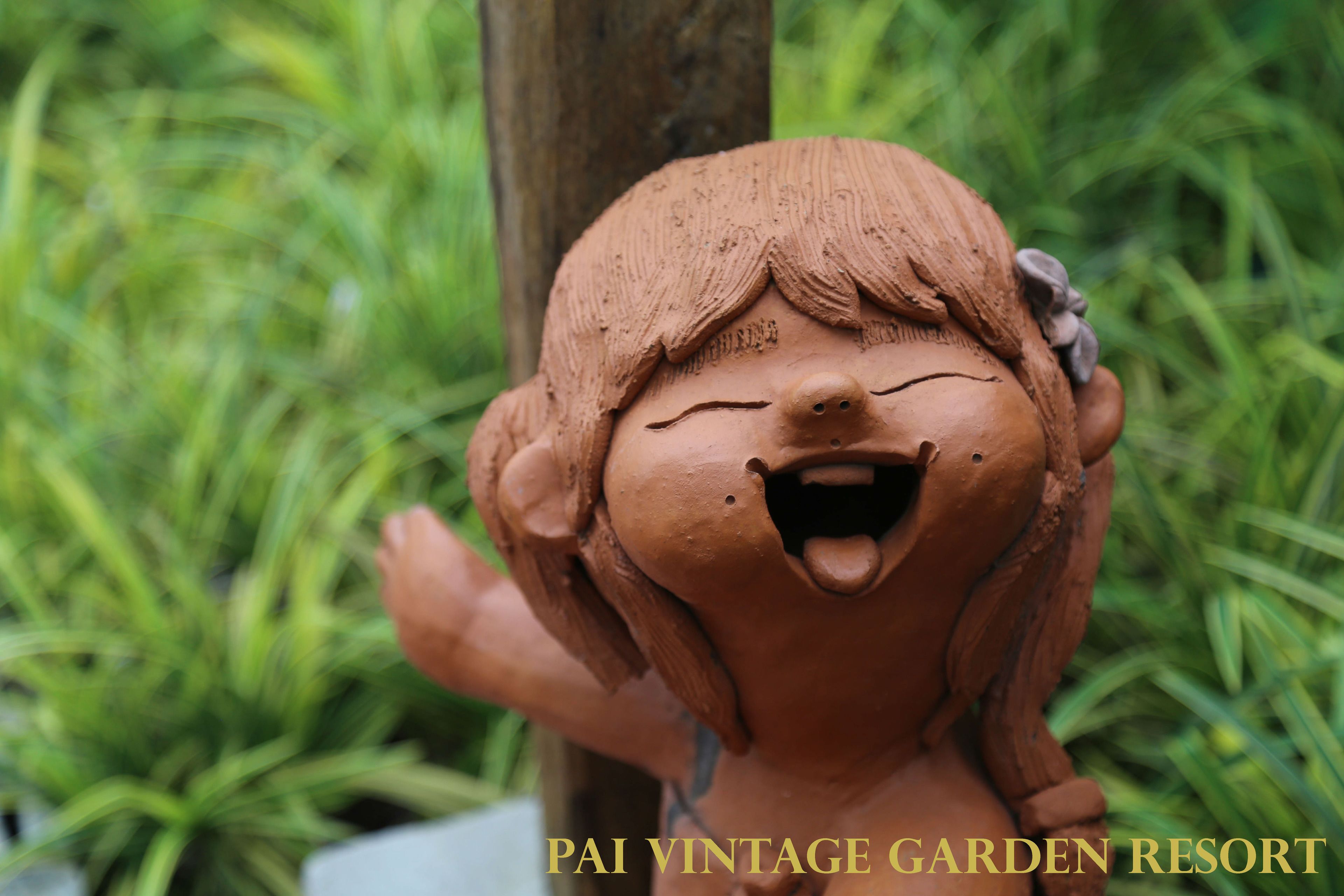 Une sculpture de jardin en terre cuite, représentant un visage souriant et orné d’une fleur dans les cheveux, le tout mis en valeur par un fond de feuillage verdoyant.