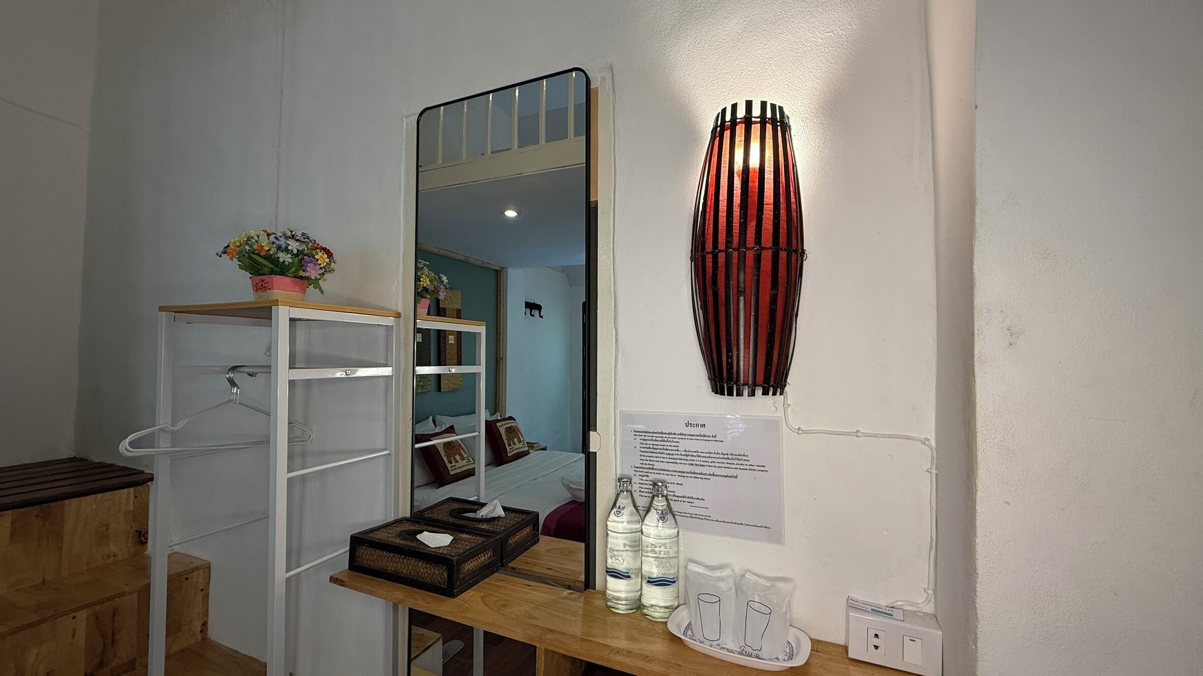 Un baño con espejo, repisa con planta en maceta, mesa de madera con botellas y vasos, y lámpara de pared.