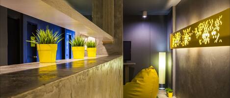 Um interior moderno com um balcão de recepção, plantas em vasos, um puff amarelo e uma parede com texto iluminado.