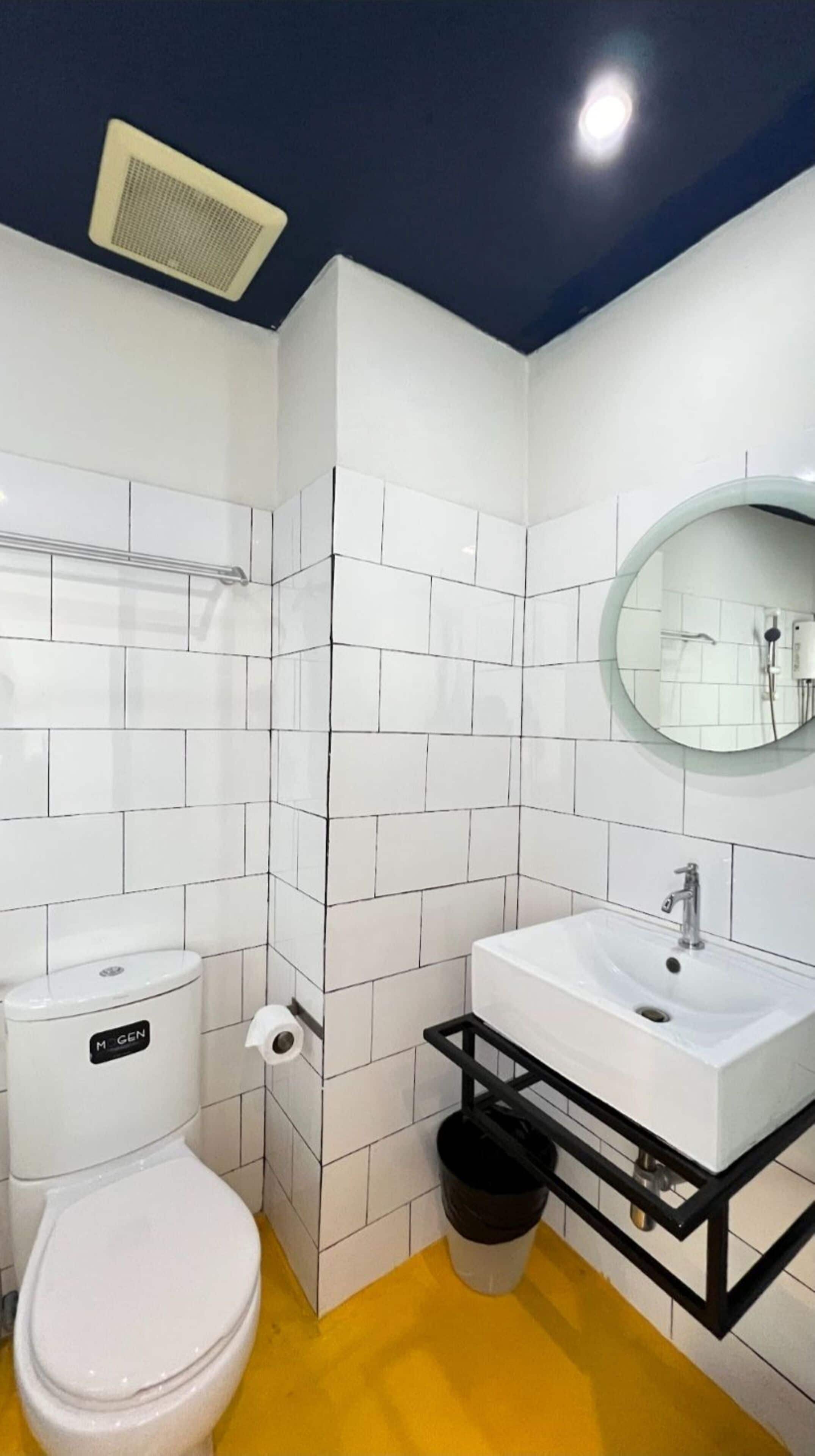 A bathroom with white tiled walls, a yellow floor, a white toilet, a white sink with a black frame, and a round mirror.