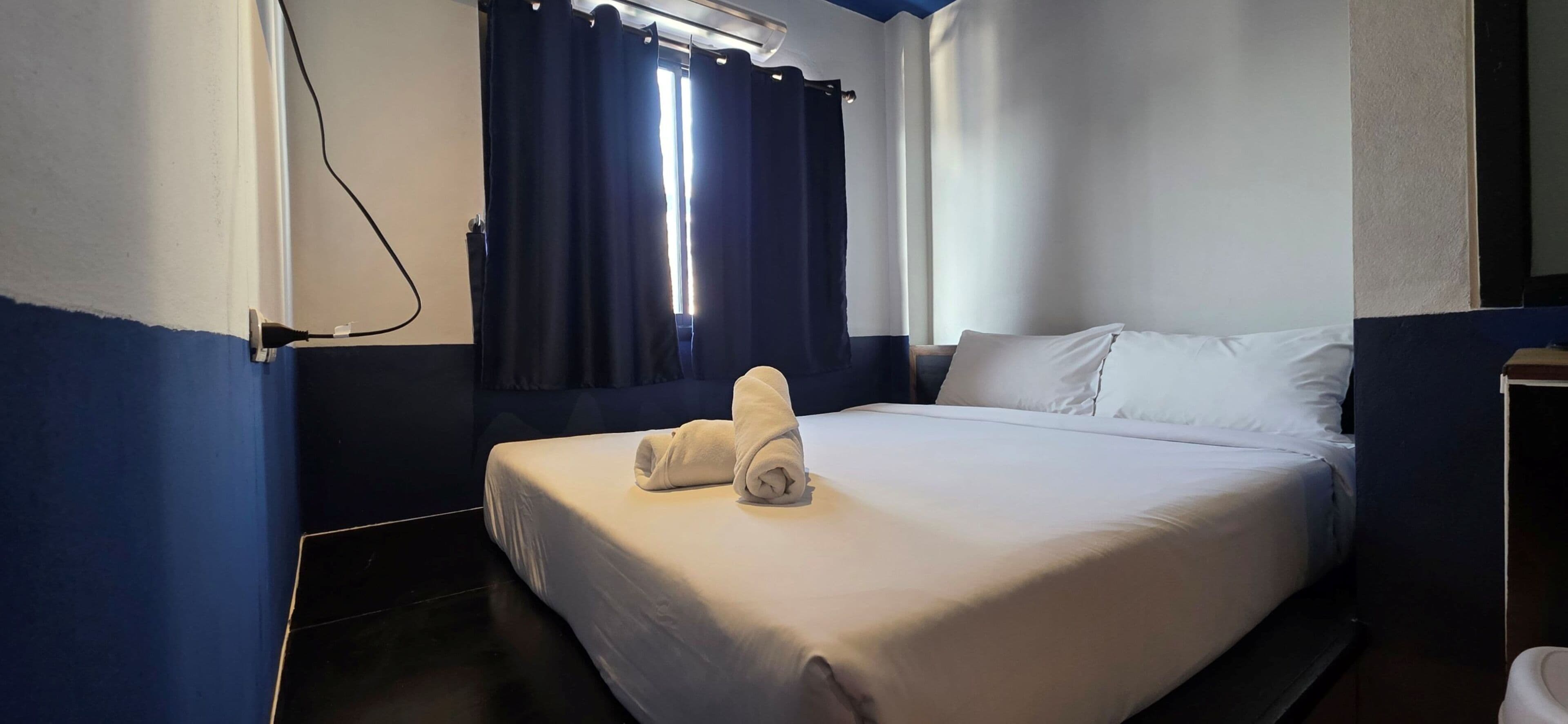 Quarto de hotel com cama, janela com cortinas, tomada e uma pequena mesa.