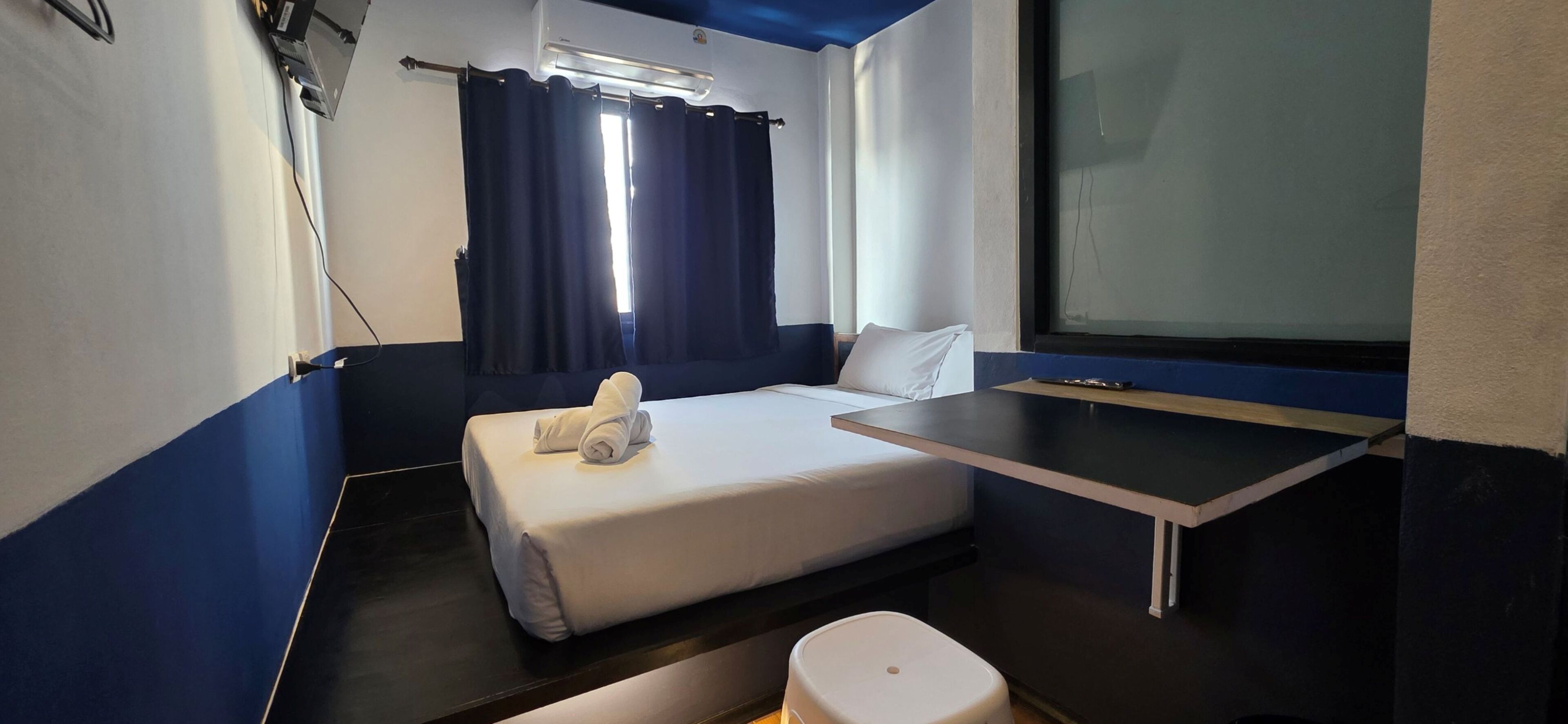 A compact hotel room with a bed, a small table, a chair, a window with curtains, and an air conditioning unit.