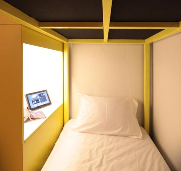 Una habitación pequeña con cama individual, una ventana a un lado, una tablet sobre el alféizar y una pared blanca lisa.