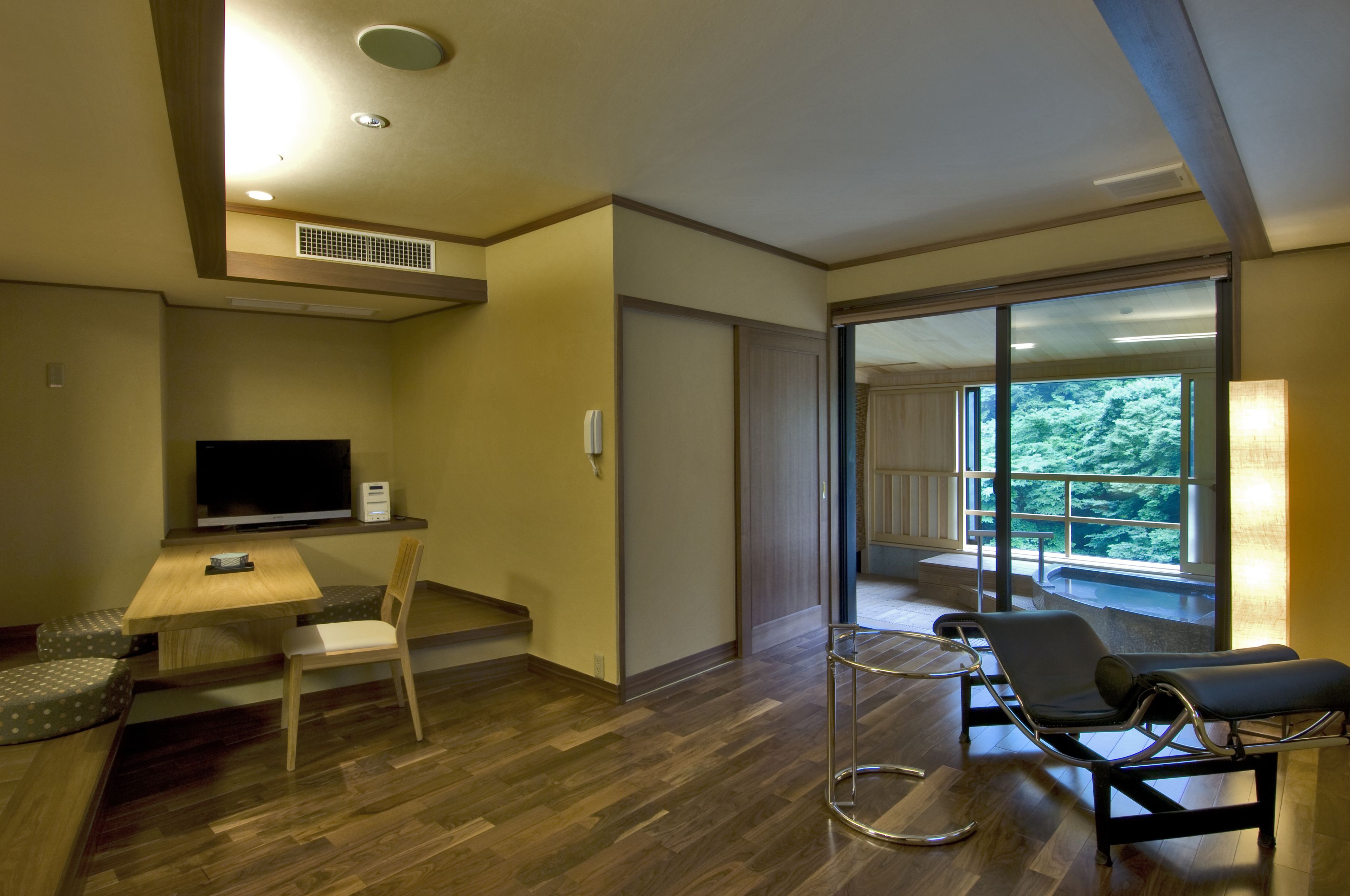 A modern hotel room with a wooden floor, a desk with a chair, a TV, and a large window offering a view of the outdoors.