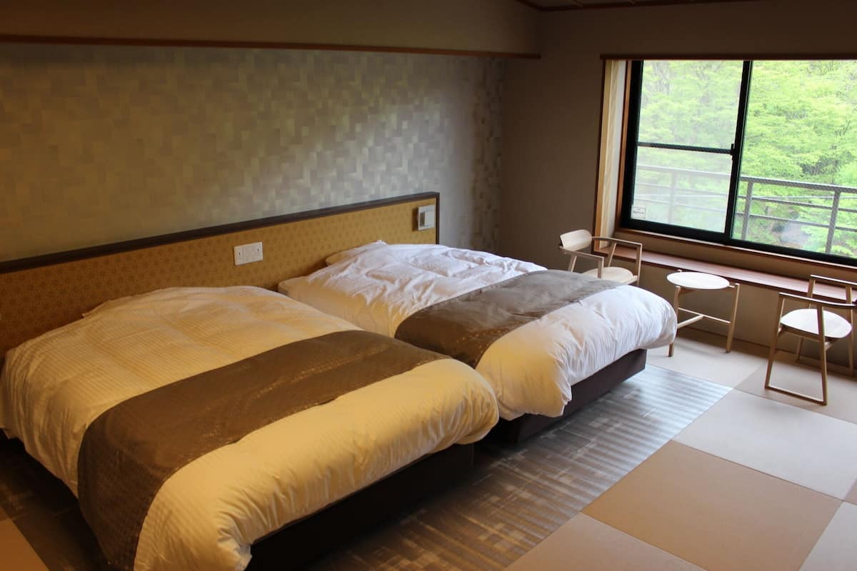 A room with two beds, a small table, and a window with a view of greenery.