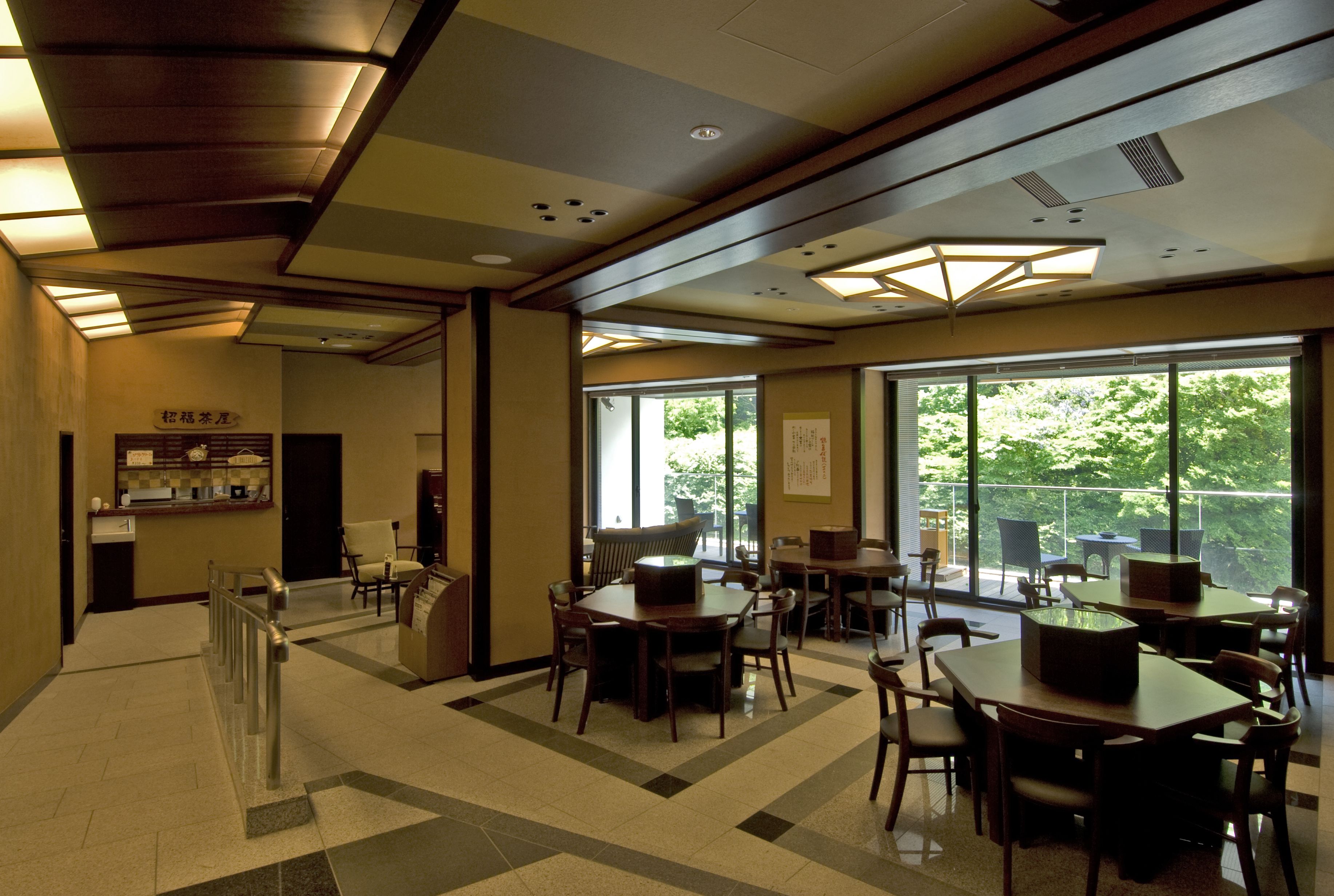 A spacious indoor area with multiple tables and chairs, large windows offering a view of greenery outside, and a reception desk with a counter.