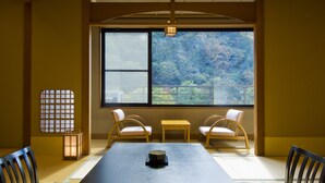 A traditional Japanese room with tatami flooring, a low table, two cushioned chairs, a sliding door with a view of a forest, and a lantern.
