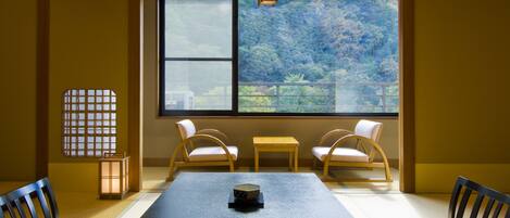 A traditional Japanese room with tatami flooring, a low table, two cushioned chairs, a sliding door with a view of a forest, and a lantern.