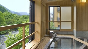 A traditional Japanese onsen with a wooden interior, a stone hot spring, and a view of a forested valley.