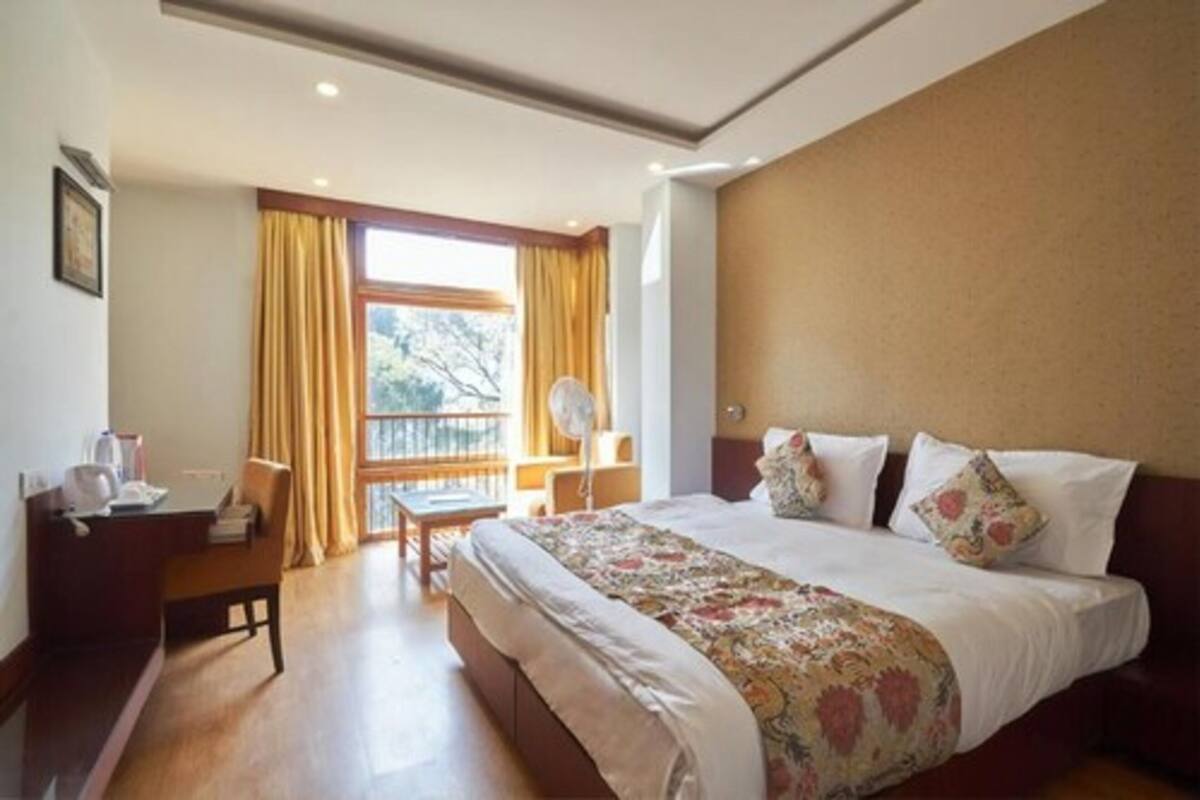 A hotel room with a large bed, a desk with a chair, a window with curtains, and a view of trees outside.