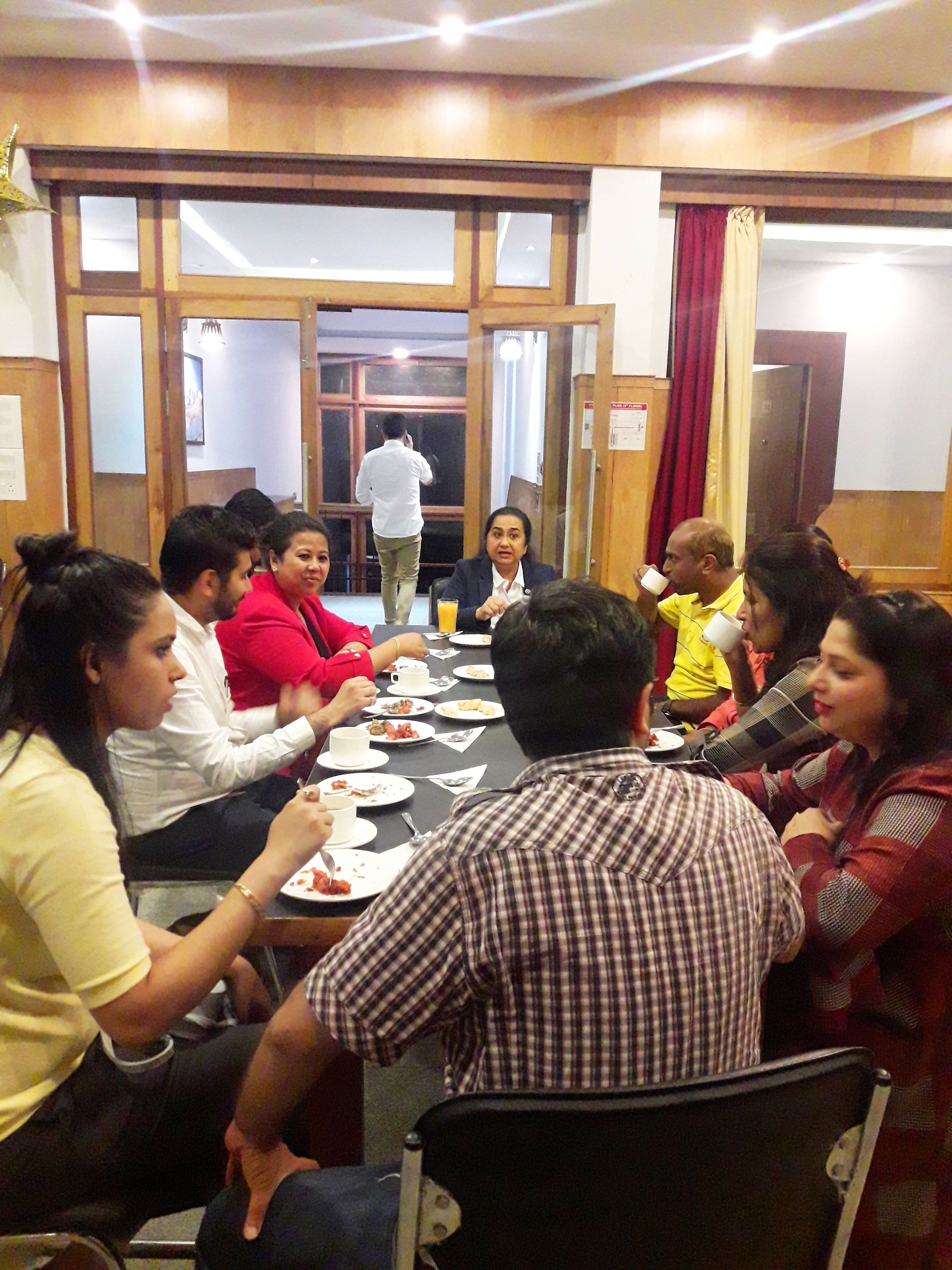 A group of people seated around a long table in a room with wooden paneling and large windows.
