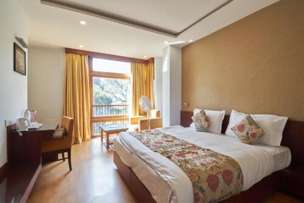 A hotel room with a bed, a desk, a chair, a window with curtains, and a view of trees.