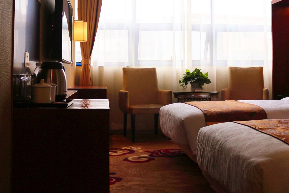 A hotel room with a bed, two chairs, a desk with a lamp, a coffee maker, and a window with curtains.
