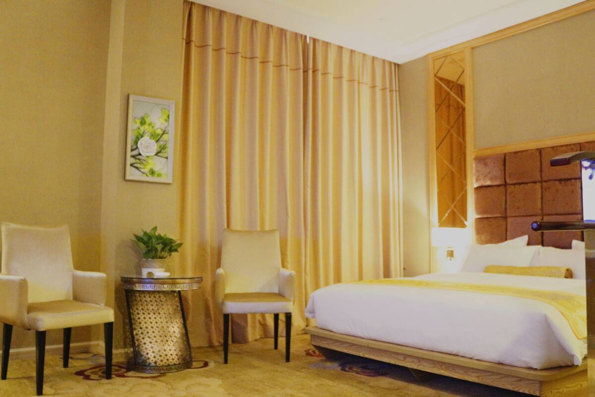 A hotel room with a bed, two chairs, a small table with a plant, a painting, and curtains.