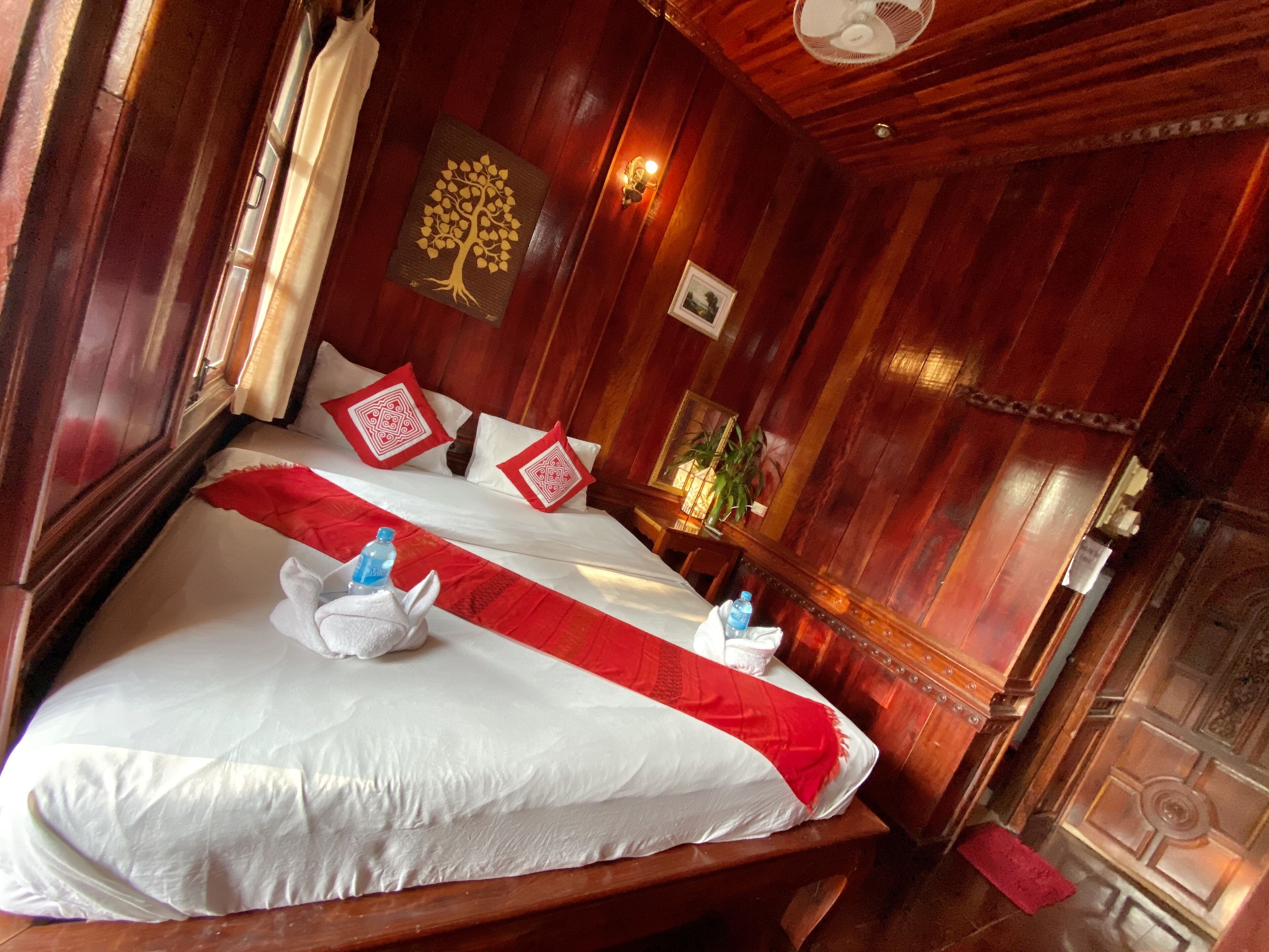 Habitación de madera con cama, almohadas, un ventilador de pared y una pequeña mesa con una botella de agua.