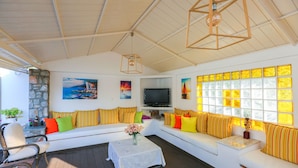 A sunlit living room with a white sofa, colorful cushions, a TV, and a stone wall. - Skiathos Island Suites (Skiathos)