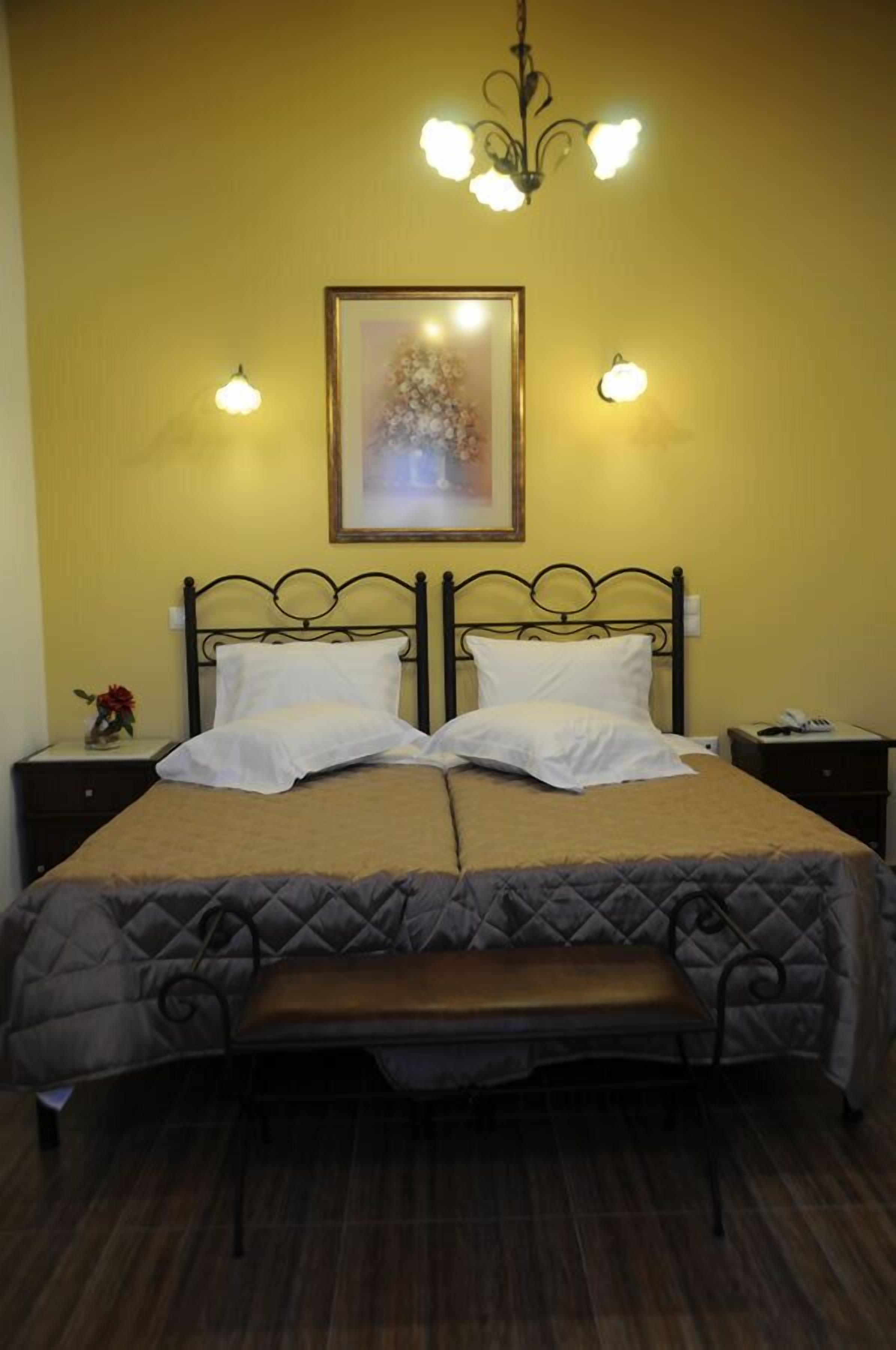 A double bed with a quilted headboard, two bedside tables, a bench, a framed picture on the wall, and a chandelier.