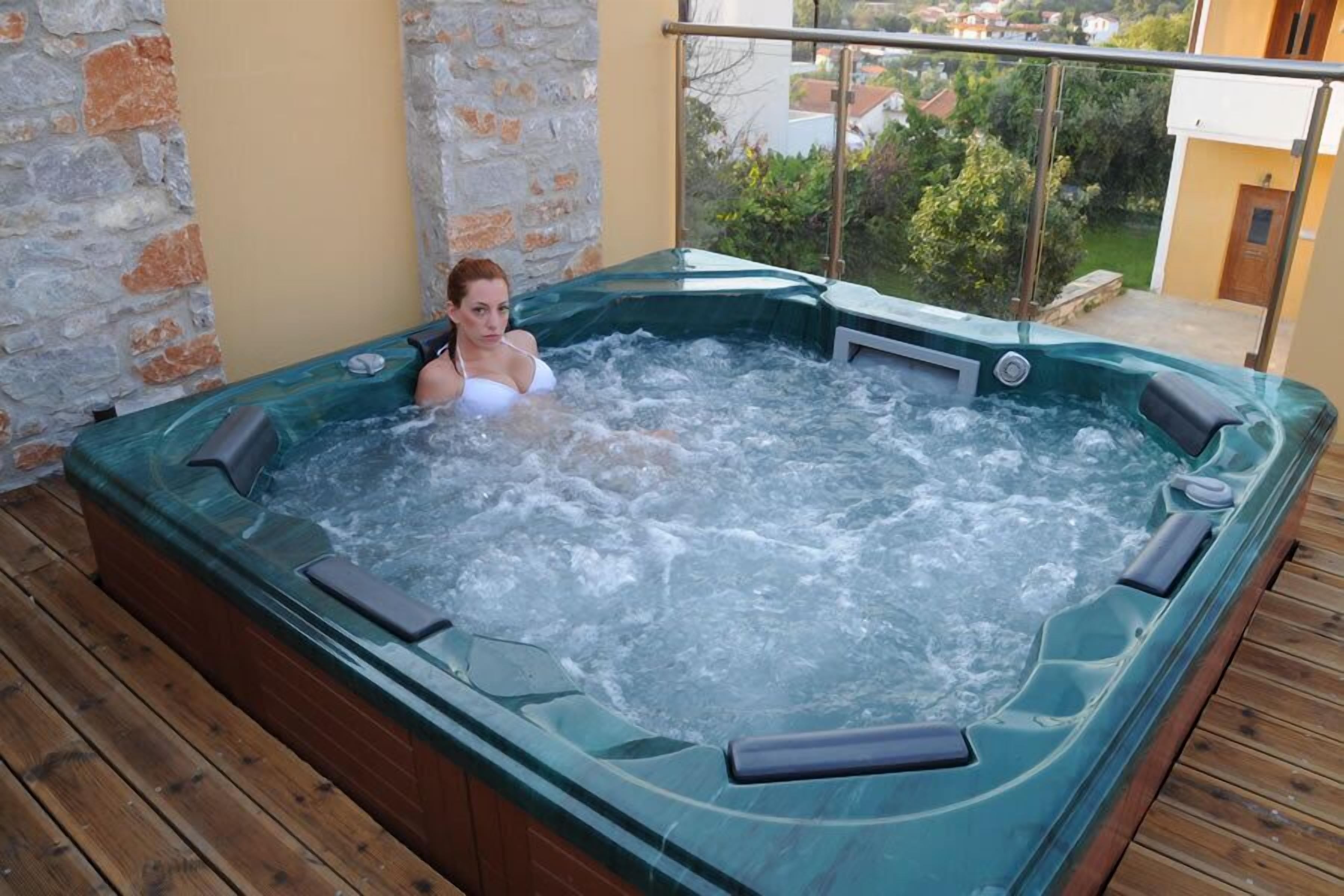 A hot tub on a wooden deck with a stone wall backdrop and a view of houses in the distance.