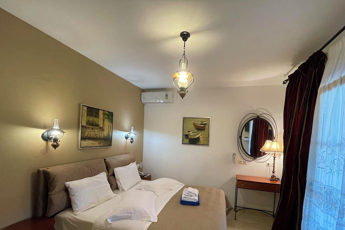 Une chambre d’hôtel avec un lit, deux lampes fixées au mur, un miroir rond, un bureau et une fenêtre avec des rideaux.