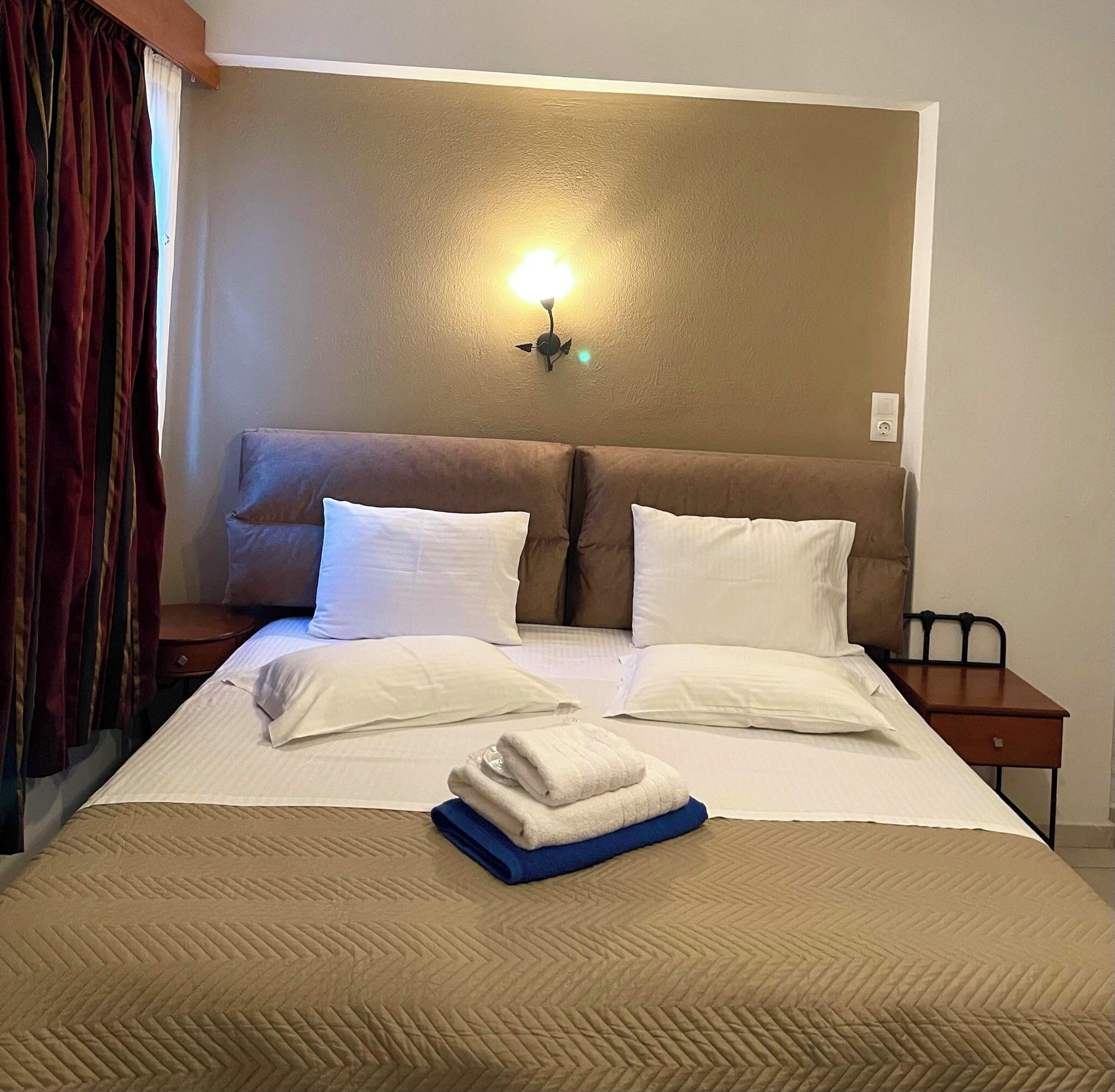 Ein Hotelzimmer mit einem großen Bett, zwei Kissen, einem Nachttisch, einer Lampe und einem Fenster mit Vorhängen.
