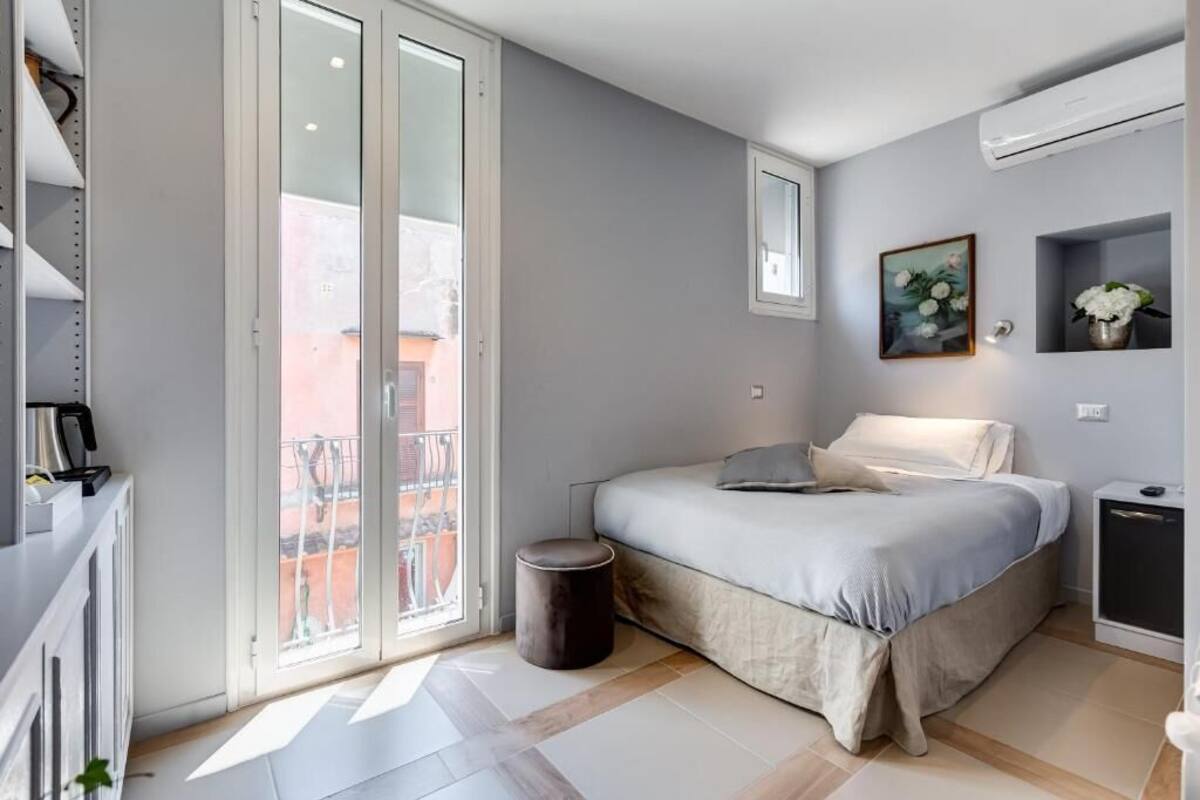 A modern bedroom with a bed, bedside table, and a small stool. There is a sliding glass door leading to an outdoor area.