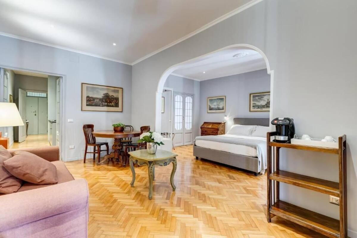 A spacious room with a wooden herringbone floor, a bed, a sofa, a dining table with chairs, and a coffee machine.