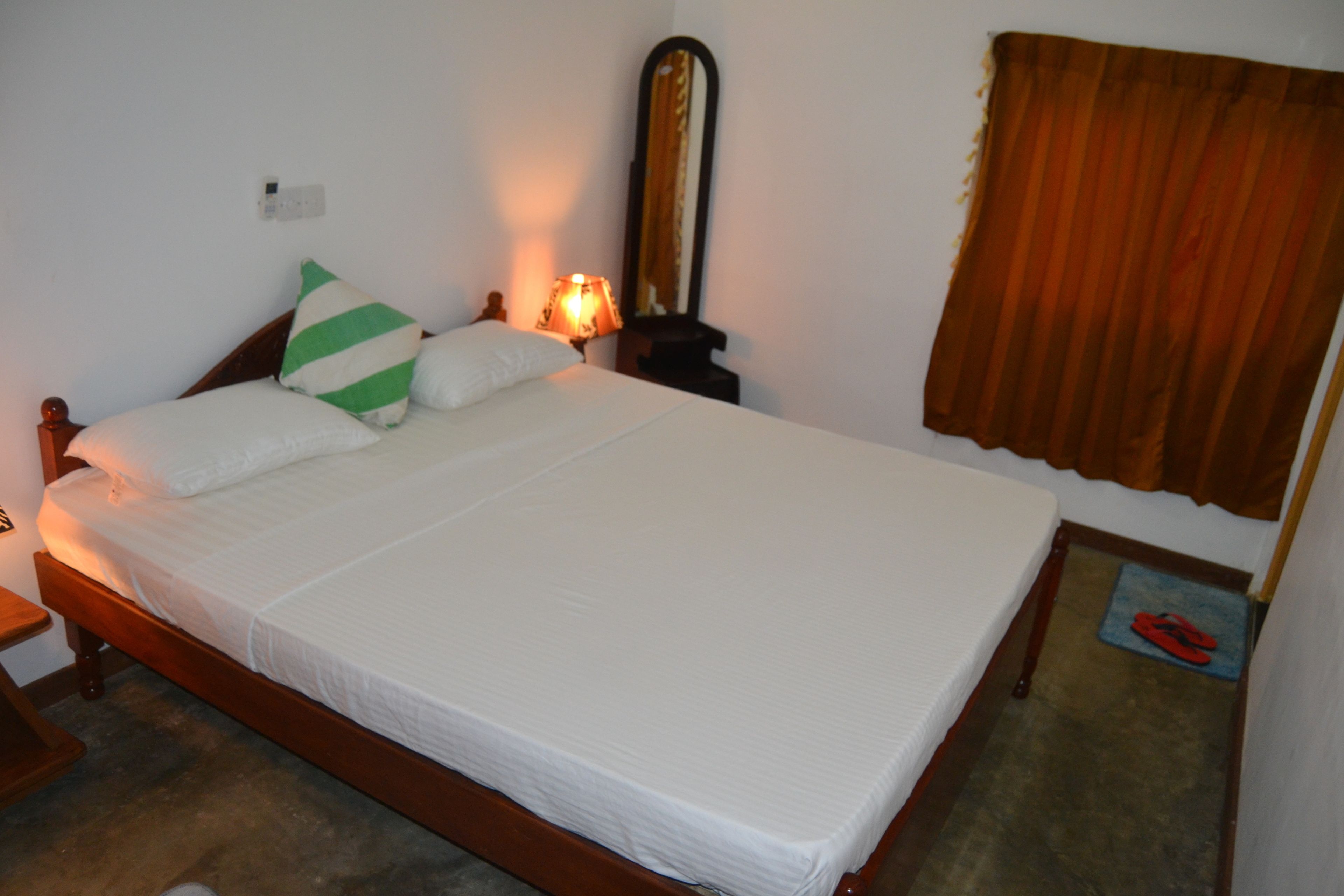 Una cama bien hecha con sábanas blancas y un cojín a rayas verdes, una lámpara de noche, un espejo y una cortina marrón.